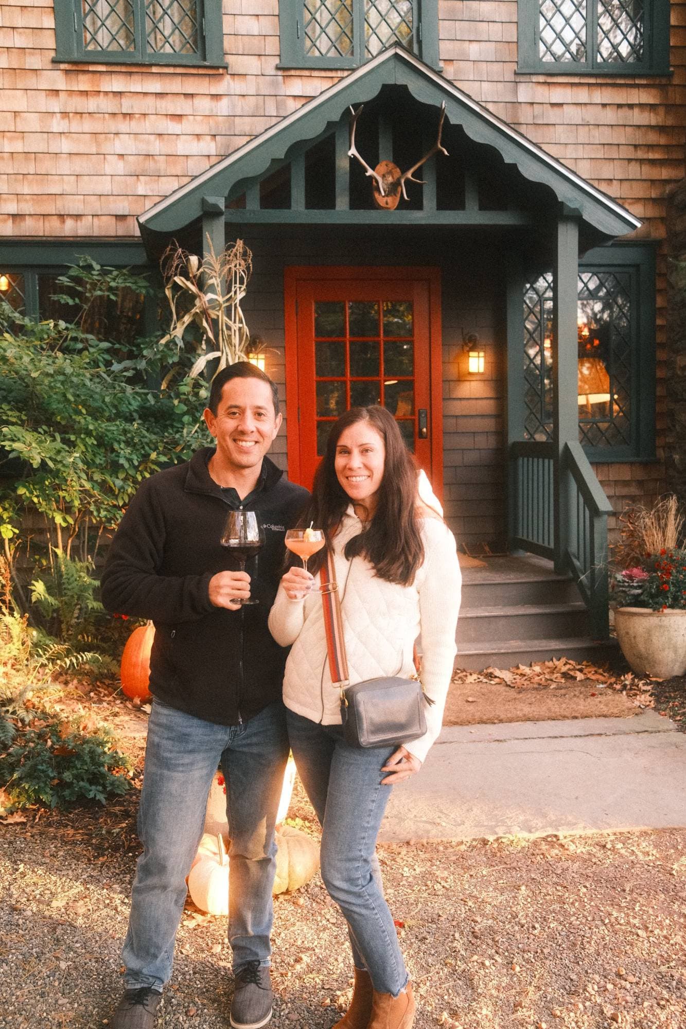 Kevin and a companion raising a glass of wine and a cocktail outside a shingled house with a red door in autumn